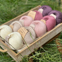 10 balls of yarn are sat in a wooden tray on a stool, sitting in grass. There are two balls of each colour. The colours from left to right are Natural, Wicker, Althaea, Bramble and Black Tulip. The yarns create a fade effect from natural cream, through pink to dusky purple.