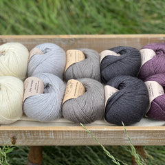 10 balls of yarn are sat in a wooden tray surrounded by grass. There are two balls of each colour. The colours from left to right are Natural, Rain, Steel, Charcoal and Black Tulip. The yarns create a fade effect from natural cream, through shades of grey ending with an accent of dusky purple.