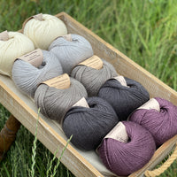 10 balls of yarn are sat in a wooden tray surrounded by grass. There are two balls of each colour. The colours from top to bottom are Natural, Rain, Steel, Charcoal and Black Tulip. The yarns create a fade effect from natural cream, through shades of grey ending with an accent of dusky purple.