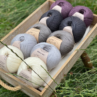 10 balls of yarn are sat in a wooden tray surrounded by grass. There are two balls of each colour. The colours from bottom left to top right are Natural, Rain, Steel, Charcoal and Black Tulip. The yarns create a fade effect from natural cream, through shades of grey ending with an accent of dusky purple.