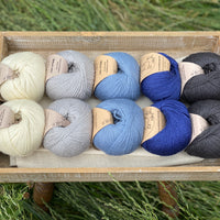 10 balls of yarn are sat in a wooden tray surrounded by grass. There are two balls of each colour. The colours from left to right are Natural, Rain, Estuary, Night Sky and Charcoal. The yarns create a fade effect from natural cream through shades of blue, ending in dark grey.