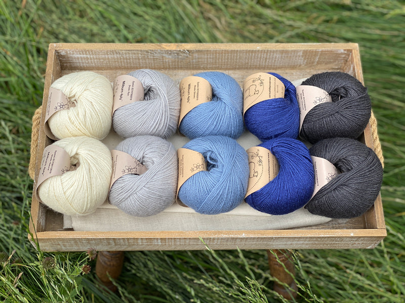 10 balls of yarn are sat in a wooden tray surrounded by grass. There are two balls of each colour. The colours from left to right are Natural, Rain, Estuary, Night Sky and Charcoal. The yarns create a fade effect from natural cream through shades of blue, ending in dark grey.