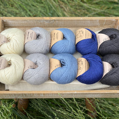 10 balls of yarn are sat in a wooden tray surrounded by grass. There are two balls of each colour. The colours from left to right are Natural, Rain, Estuary, Night Sky and Charcoal. The yarns create a fade effect from natural cream through shades of blue, ending in dark grey.