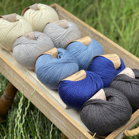 10 balls of yarn are sat in a wooden tray surrounded by grass. There are two balls of each colour. The colours from top left to bottom right are Natural, Rain, Estuary, Night Sky and Charcoal. The yarns create a fade effect from natural cream through shades of blue, ending in dark grey.