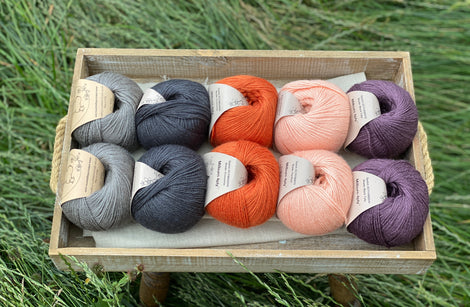 10 balls of yarn are sat in a wooden tray surrounded by grass. There are two balls of each colour. The colours from left to right are Steel, Charcoal, Crocosmia, Tea Rose and Black Tulip. The yarns create a palette of greys with orange, peach and dusky purple.