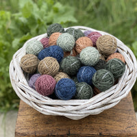 A small round white wicker basket containing Yarnlings (small balls of yarn) in a variety of colours including blue, grey, green, brown, pink and purple. The basket sits on a wooden stool with green foliage in the background.