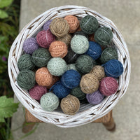 A small round white wicker basket containing Yarnlings (small balls of yarn) in a variety of colours including blue, grey, green, brown, pink and purple. The basket sits on a wooden stool on concrete paving..