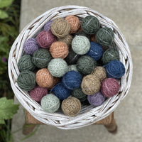 A small round white wicker basket containing Yarnlings (small balls of yarn) in a variety of colours including blue, grey, green, brown, pink and purple. The basket sits on a wooden stool on concrete paving..