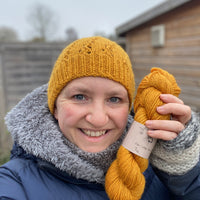 Victoria wearing a hat knitted in the same rich yellow colourway as the skein in her hand