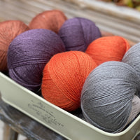 Eight balls of yarn in four pairs. From left to right the colourways are red-brown, purple, orange and grey