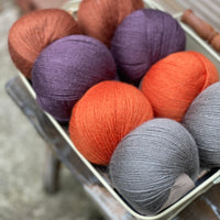 Eight balls of yarn in four pairs. From left to right the colourways are red-brown, purple, orange and grey