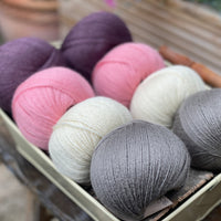 Eight balls of yarn in four pairs. From left to right the colourways are purple, pink, cream and grey