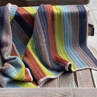 A striped crochet blanket draped over a bench. The stripes are rust, green, blue, brown and black and the blanket has a beige edging