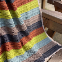 A striped crochet blanket draped over a bench. The stripes are rust, green, blue, brown and black and the blanket has a beige edging