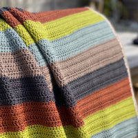 A striped crochet blanket draped over a bench. The stripes are rust, green, blue, brown and black and the blanket has a beige edging